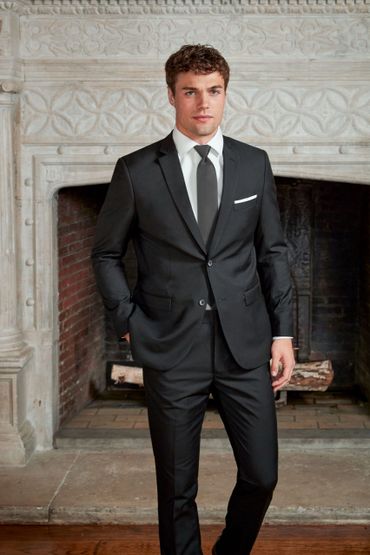 Young man in a black suit standing confidently in front of an ornate fireplace.