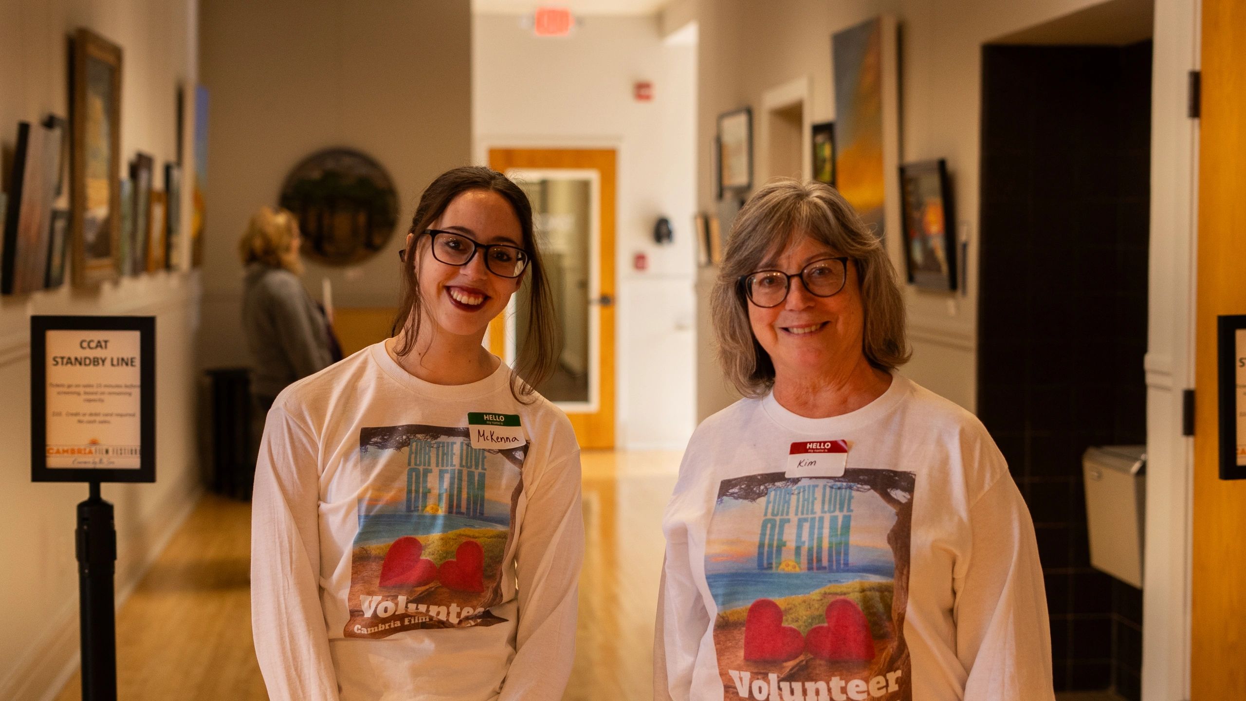 Two smiling volunteers wearing matching shirts at an indoor event.
