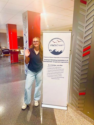 VP of Recruitment, Ryleigh Horn, waving and standing outside of AMA's meeting by a sign with their l