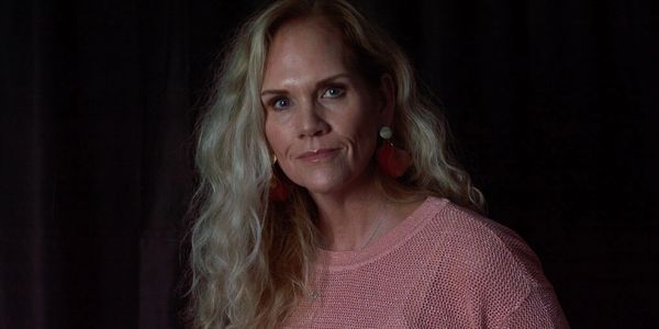 Woman with blonde curly hair wearing a pink sweater and floral earrings against a dark background.