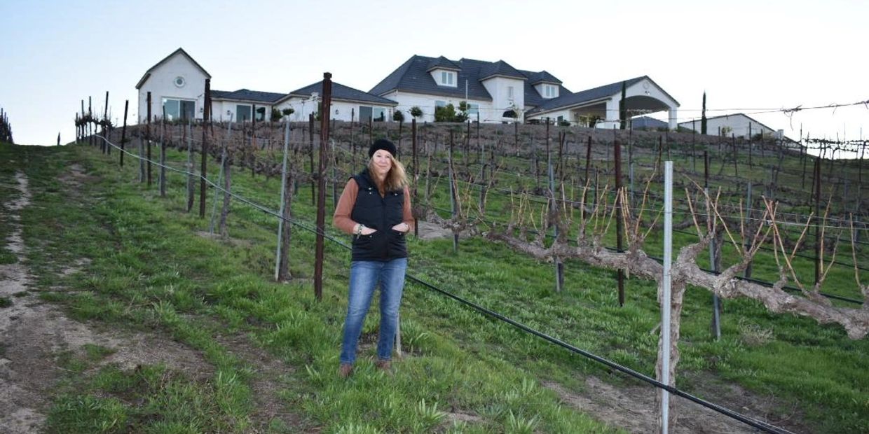 A woman stands in a vineyard with a large house in the background.