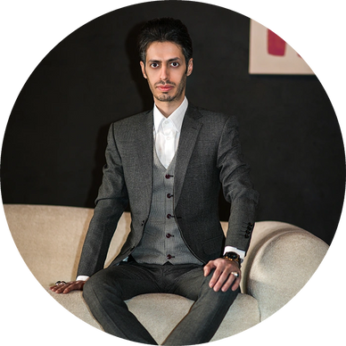 Man in a sharp gray suit sitting on a beige couch against a dark background.