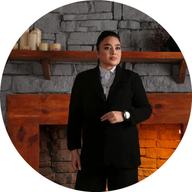 Confident person in a black suit standing by a stone fireplace.