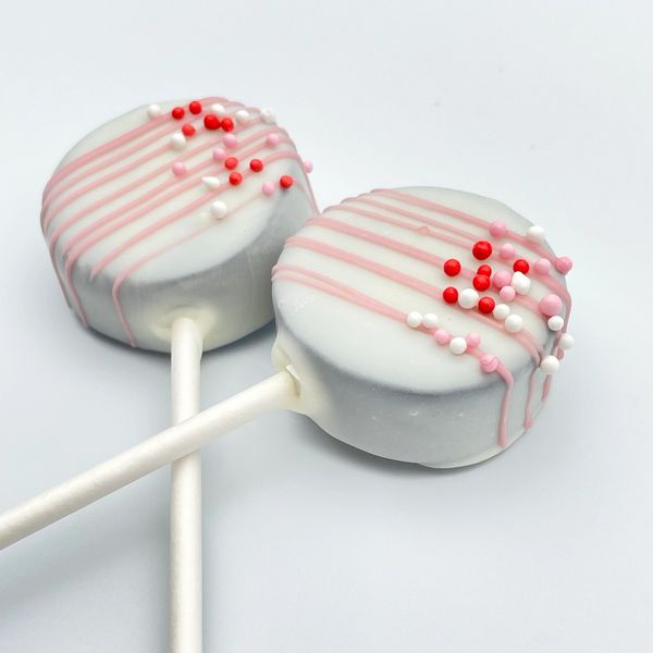 Two white chocolate cake pops with pink drizzle and colorful sprinkles.