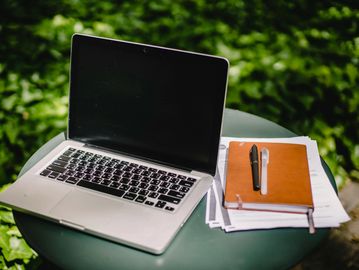 A laptop and notebook with pens on a round table outdoors.