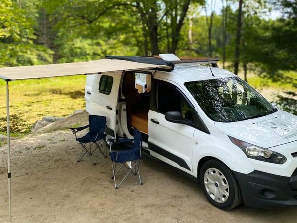 White camper van with awning and two camping chairs in a forest setting.