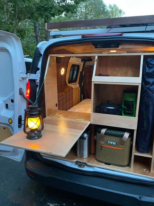 Van converted into a cozy camping setup with wooden shelves and a lantern.