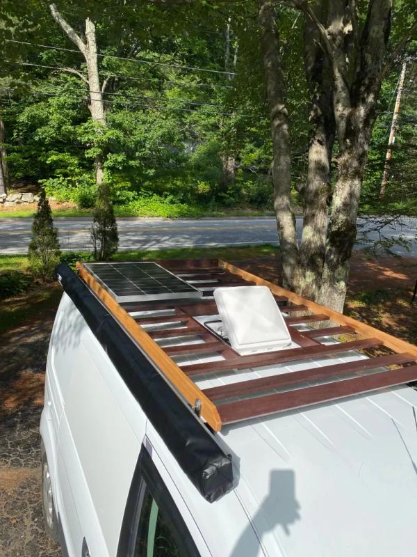 White van with wooden roof rack and solar panel under trees.