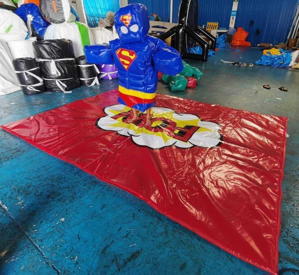 Child wearing inflatable Superman suit on a red mat indoors.