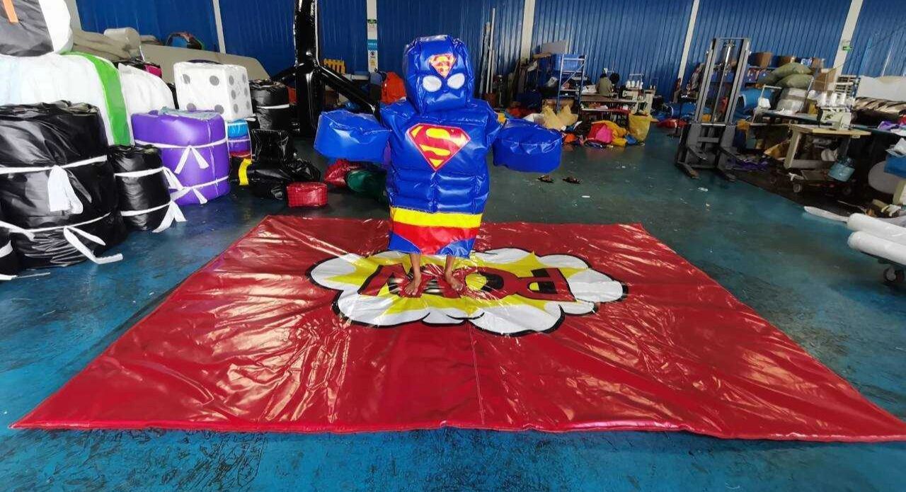 Person in inflatable Superman suit stands on red mat indoors.
