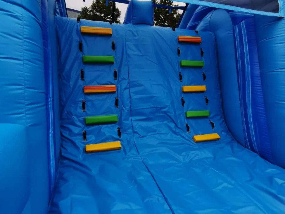 Blue inflatable climbing wall with colorful handholds outdoors.