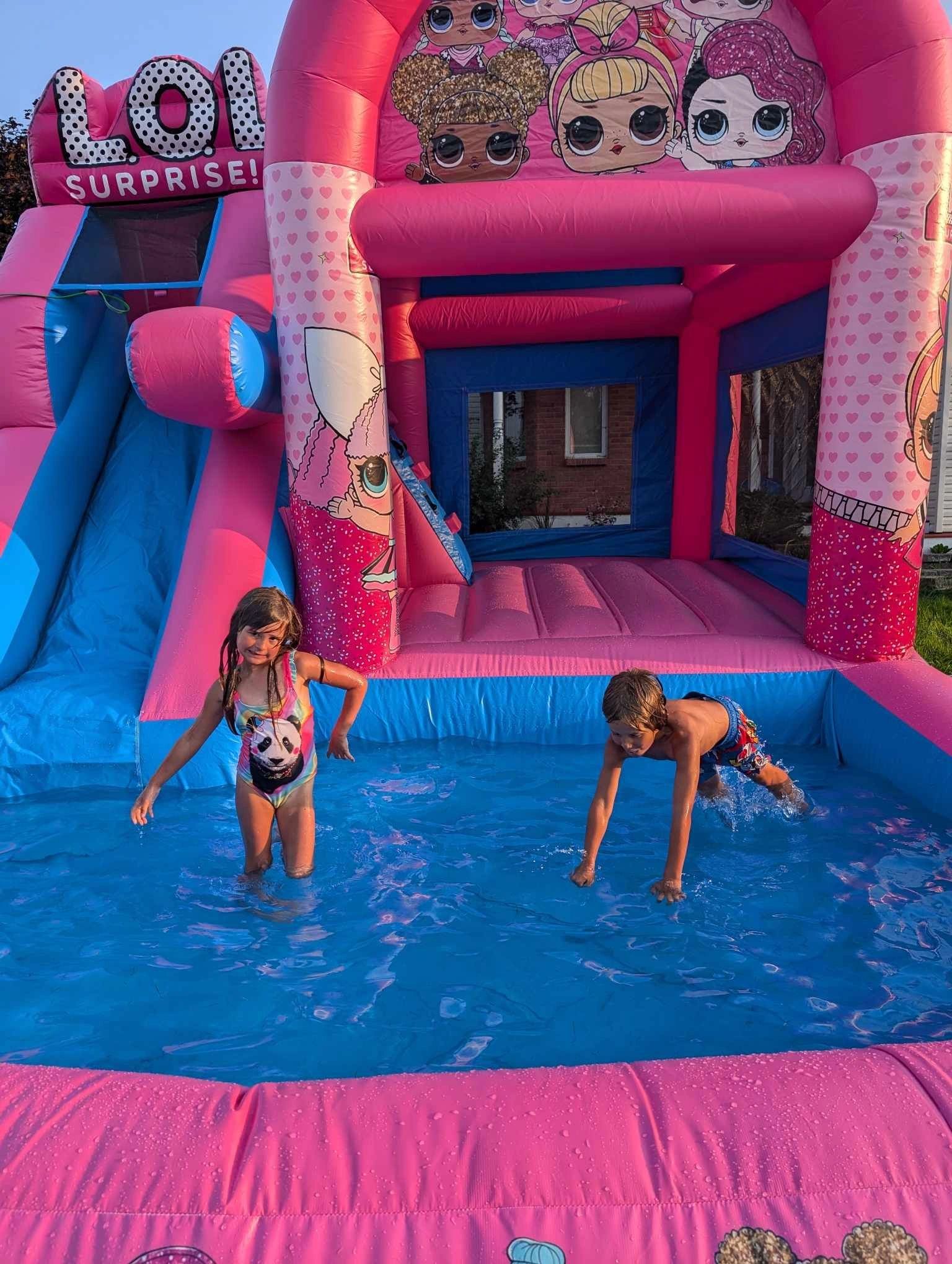 Two children playing in a pink and blue LOL Surprise inflatable water slide.