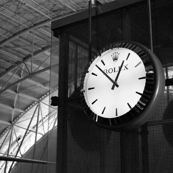 Black and white image of a Rolex clock showing 12:06 inside a building with a high ceiling.