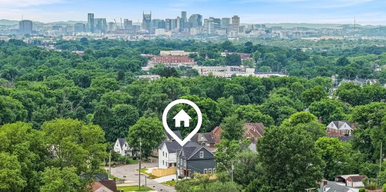 A suburban neighborhood with dense trees and a city skyline in the background under a blue sky.