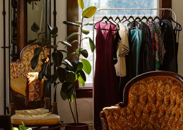 A cozy room with vintage chairs, a plant, and a rack of colorful dresses by the window.