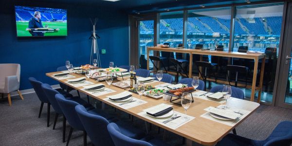 A dining table set in a stadium lounge with a sports broadcast on TV.