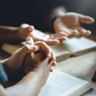 Hands clasped in prayer with open hands and books in the background.