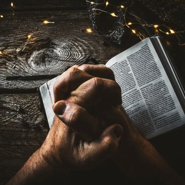 Hands clasped in prayer over an open Bible with warm fairy lights nearby.