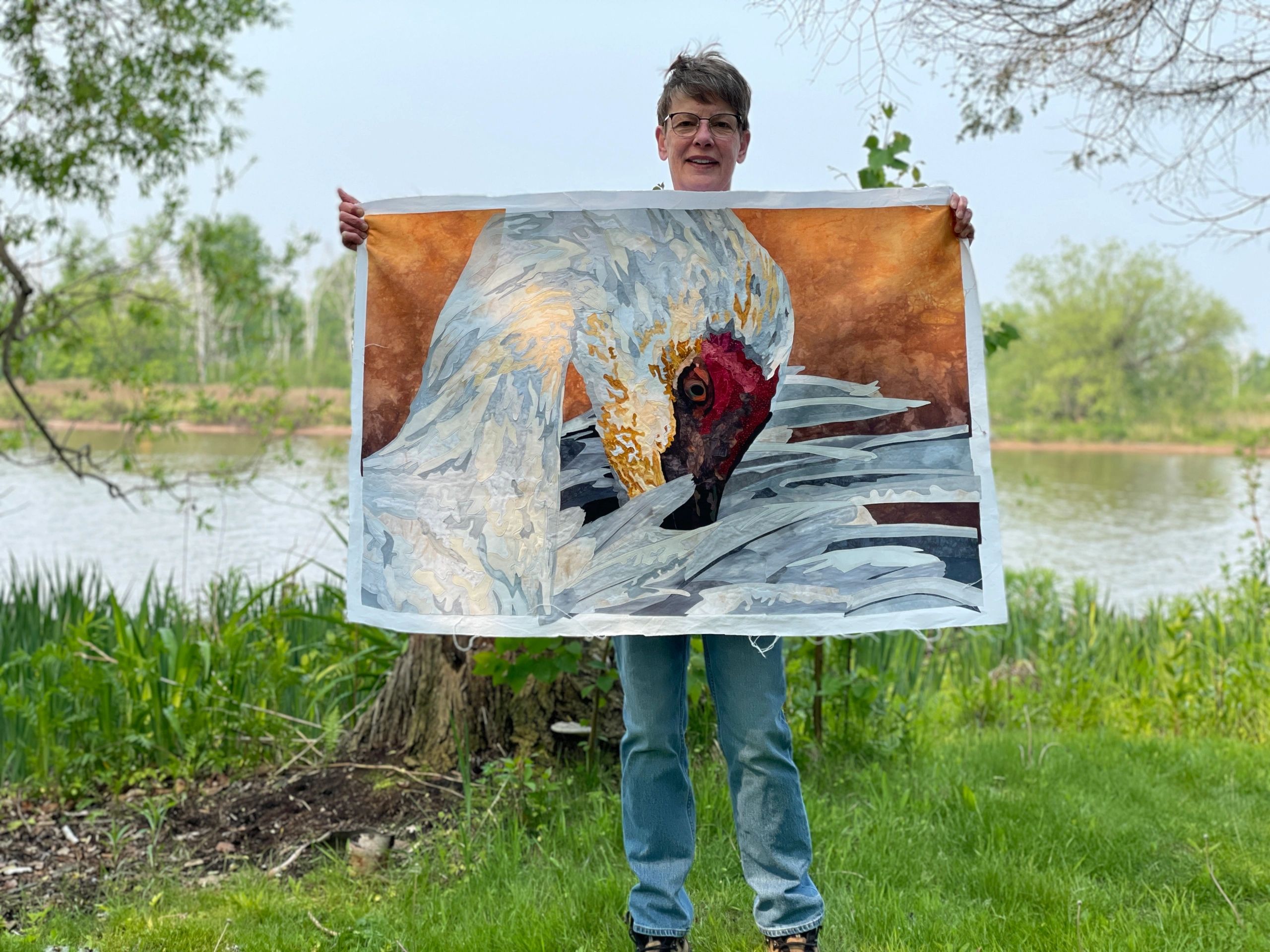A woman holding a Sandhill Crane fabric collage with green trees and water in the background.