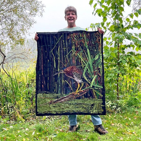 Artist holding the completed fabric collage of a Green Heron in a natural setting.