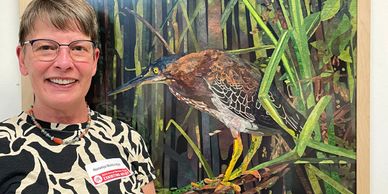 A smiling woman with glasses and short brown hair standing in front of a Green Heron fabric collage