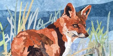 Fabric collage of a fox standing in dune grass with blue water and sky in the background
