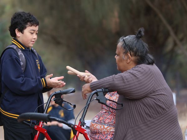An elderly woman hands drumsticks to a young boy outdoors.