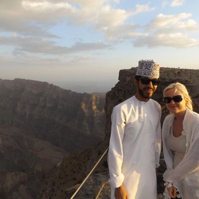 A man and woman wearing sunglasses stand on a rocky cliff with mountains in the background.