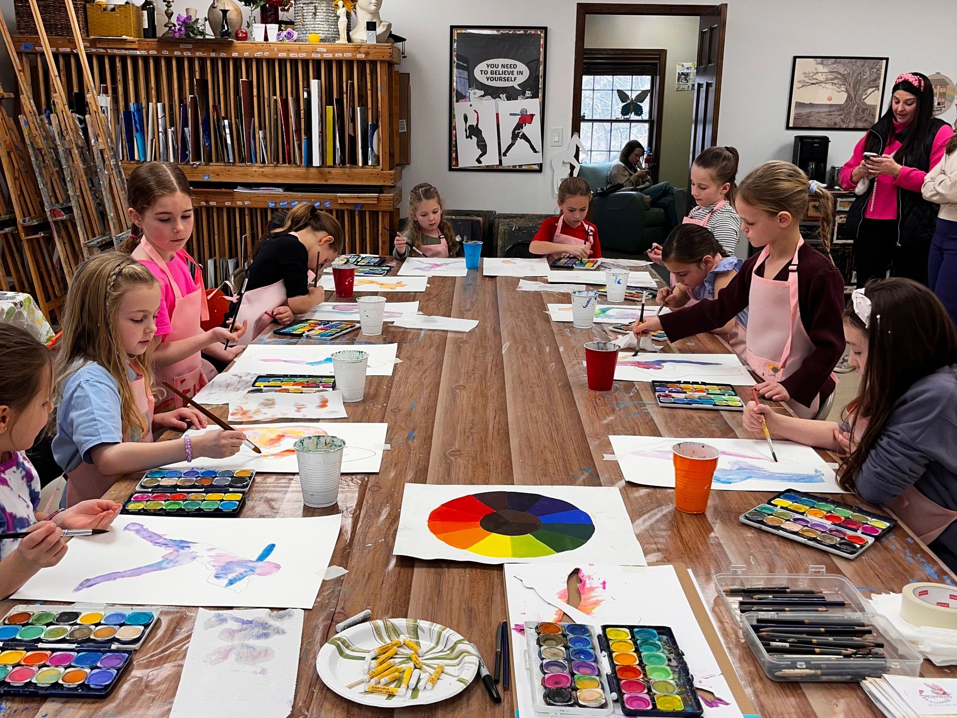 Children painting with watercolors in an art classroom around a large wooden table.