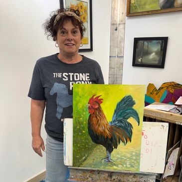 Woman proudly displays a colorful rooster painting in an art studio.