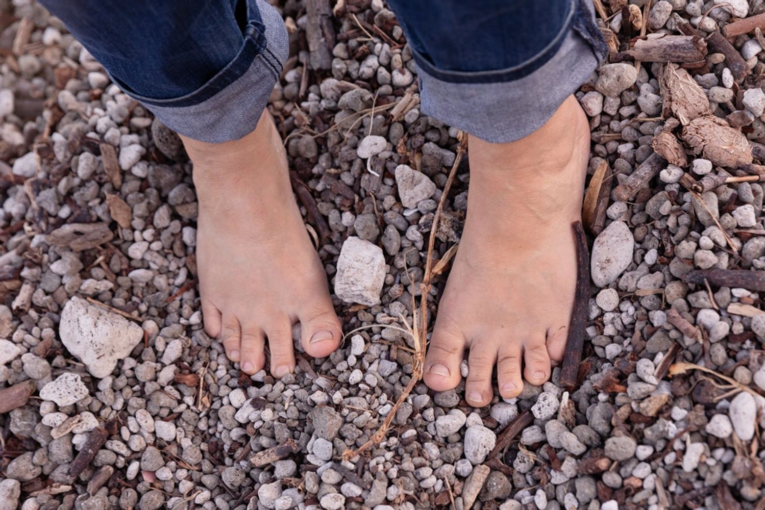 Bare feet standing on small rocks and pebbles with rolled-up jeans.