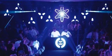 A lively nightclub scene with DJs and dancing crowd under blue lights.