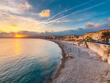 Sunset over a calm sea and pebble beach with coastal buildings.