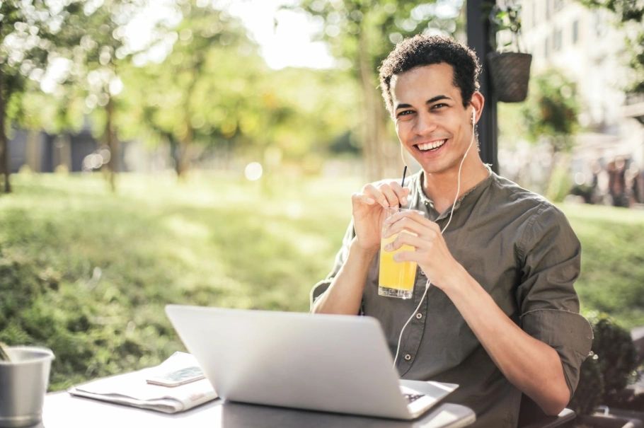 Smiling young man sitting at an outside table in front a laptop, with headphones and a cool drink.