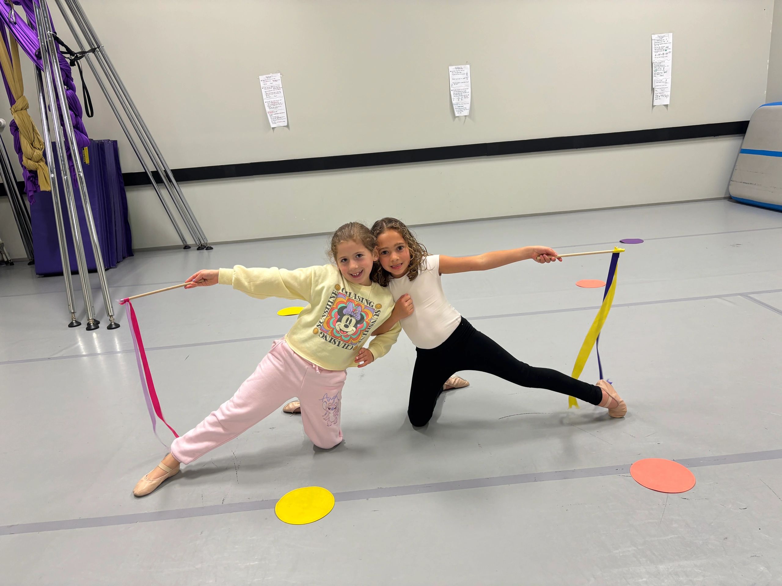 Two girls posing with ribbon sticks in a dance studio.