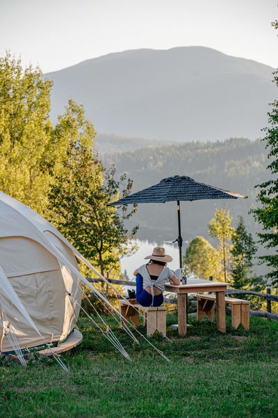 Woman seated at a picnic table under an umbrella located beside a canvas tent with the mountains and