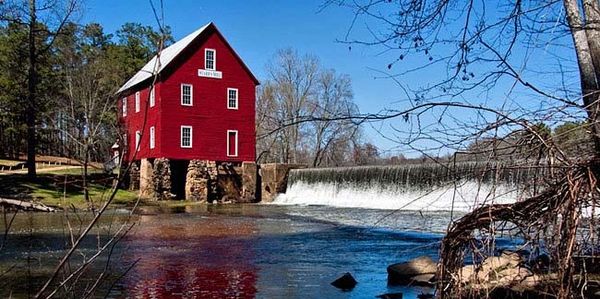 Red historic mill by flowing river and small waterfall under clear blue sky.