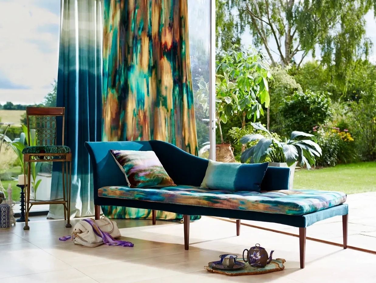 Elegant teal chaise lounge with colorful cushions by large windows overlooking a green garden.