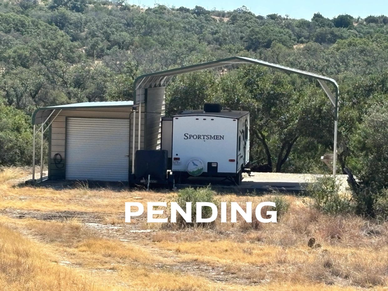 Sportsmen RV parked under a metal carport near trees.