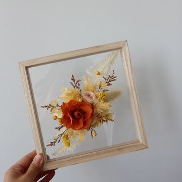 Hand holding a wooden frame with dried flowers inside against a plain wall.