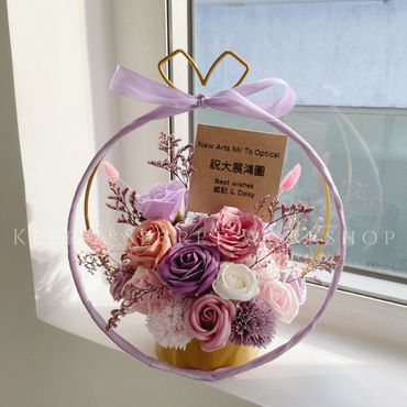 Elegant floral arrangement with pastel roses in a heart-shaped gold frame adorned with a lavender ribbon.