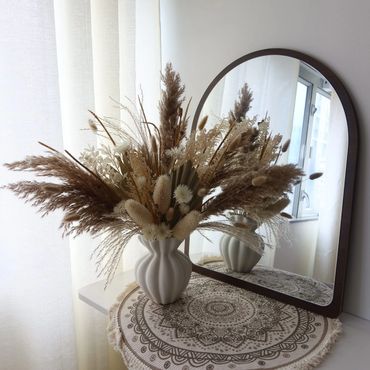 Elegant dried flower arrangement in a white vase beside an arched mirror on a decorated table.