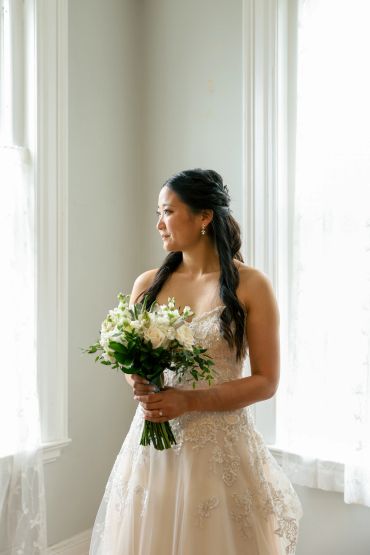 Bride in lace wedding dress holding bouquet, looking out window.