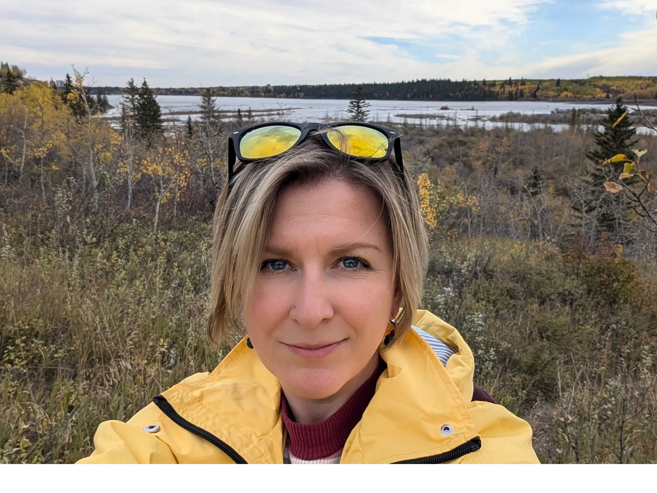 Female in a bright yellow coat in the fall, surrounded by trees, overseeing a lake in the distance.