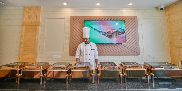 A chef stands behind empty buffet trays in a modern dining area.