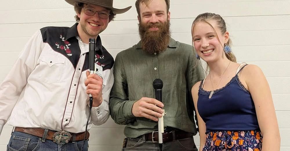 Three smiling people holding microphones, posing against a plain wall.