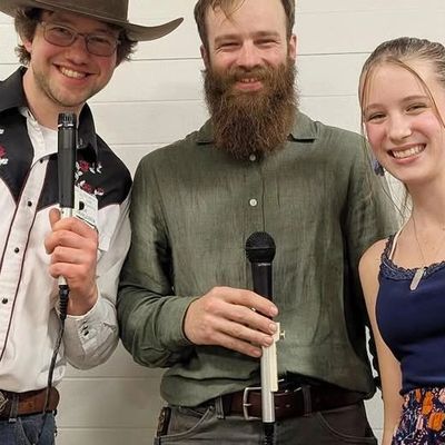 Three smiling people holding microphones, posing against a plain wall.