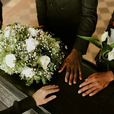 Hands of mourners resting near a casket adorned with white flowers.