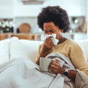 Person sneezing into tissue while holding a mug and wrapped in a blanket.