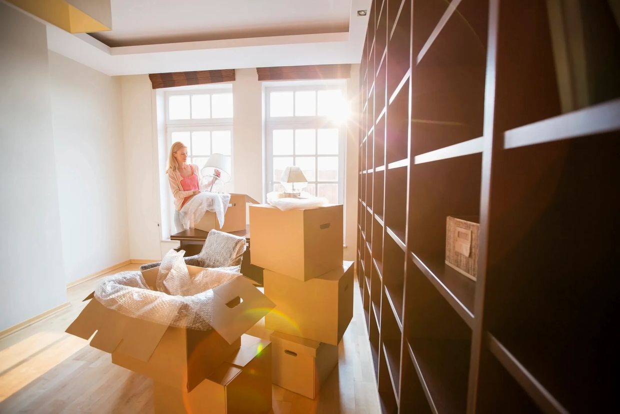 Woman unpacking a lamp and household items from moving boxes in a bright, sunlit room.